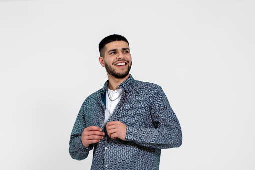 Medium portrait shot of a young Asian man taken in a studio in Newcastle Upon Tyne. He is buttoning up his shirt while smiling and looking away from the camera.\n\nVideos are available similar to this scenario.