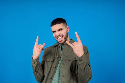 Medium portrait shot of a young Asian man taken in a studio in Newcastle Upon Tyne. He is standing looking at the camera while making rock hand gestures. He is smiling with his tongue sticking out. Taken in front of a blue background.
