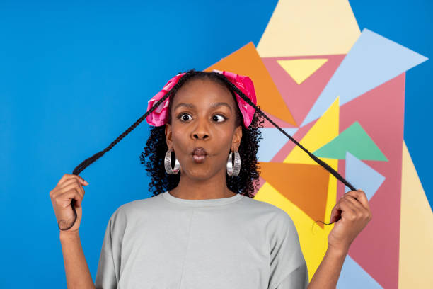 Close-up portrait shot of a young black woman in a studio in Newcastle Upon Tyne. She is pulling a funny face and holding her hair in both hands. She is standing in front of an artwork. Taken in front of a blue background.\n\nVideos are available similar to this scenario.