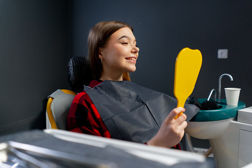 In the dental office young girl sits and looks at her teeth in a yellow dental mirror rejoicing