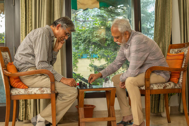 Senior male friends sitting on armchairs and playing chess with concentration while spending leisure time together at home