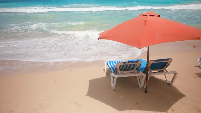 Two empty blue sun loungers under the red parasol on the sandy beach near the sea water with big waves. Rest and relaxation summer holiday scene