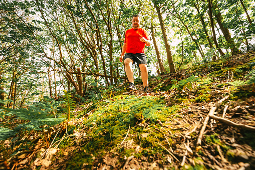 Wide angle color image depicting a man in his 30s trail running through the forest. The man is wearing sports training clothing - red t shirt, black shorts and trainers.