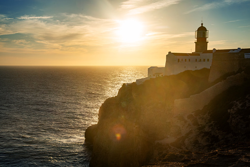 A stunning sunset paints the sky over a historic lighthouse perched atop rugged cliffs. The golden sun casts a warm glow over the Atlantic Ocean, creating a picturesque scene.