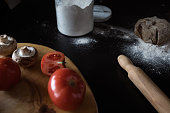 ingredients for baking pizza on dark table: tomatoes, mushrooms, rye dough, flour and rolling pin
