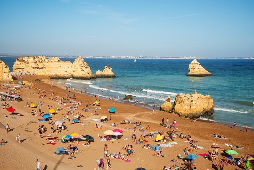 Vibrant beach scene with people enjoying a sunny day at a sandy beach in Lagos, Algarve, Portugal. Colorful umbrellas and stunning cliffs enhance the landscape.