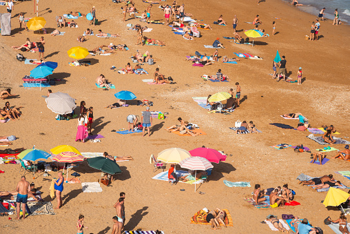 Aerial view of a bustling beach in Lagos, Algarve, Portugal filled with tourists enjoying a sunny day. Colorful umbrellas dot the sandy shore.