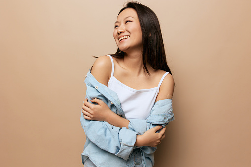 Portrait of happy tanned adorable young Asian lady after vaccination hold shirt sleeve posing isolated on beige pastel background. People Emotions. CoVID Vaccination concept. Copy Space Offer Banner.
