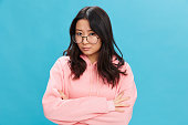 Upset cute Asian student young lady in pink hoodie sweatshirt wear round glasses cross hands posing isolated on blue studio background. The best offer for ad. Eyewear for vision correction concept