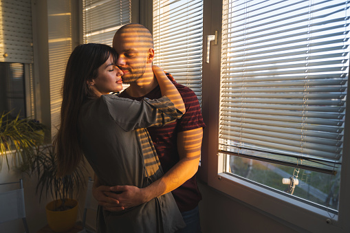 Young couple is hugging each other gently next to the window. The evening sun is shining through the blinds and shadows stripes are falling over their faces.