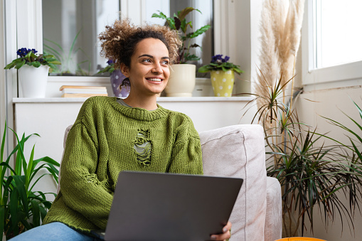 Young smiling mixed-race woman is spending her free day at home wearing a cozy green sweater, sitting in her bright living room, surfing the Internet.