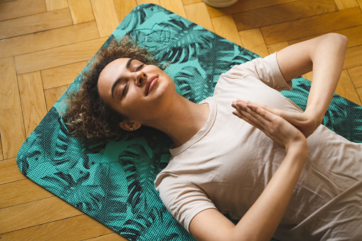 Photo from above of a mixed-raced beautiful smiling woman meditating while lying down on the yoga mat on the floor in the living room.
