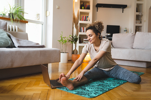 Young mixed-race woman with a curly hair is doing stretching exercises on her yoga mat in the living room while watching tutorials on the Internet.