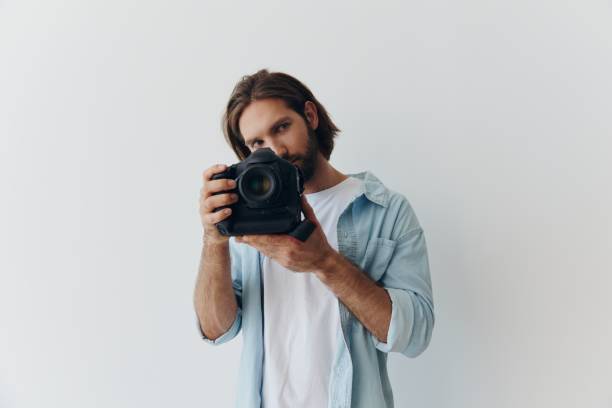 istock Man hipster photographer in a studio against a white background holding a professional camera and setting it up before shooting. Lifestyle work as a freelance photographer 2149001985