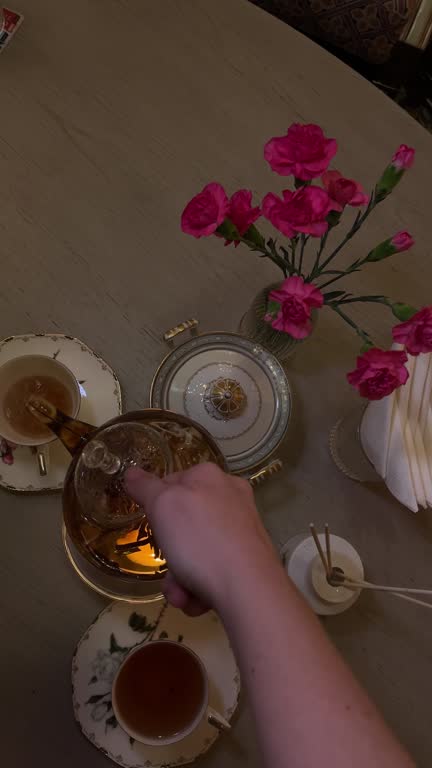 A woman's hand pours tea into a cup against a background of beautiful flowers of pink carnations Tea drinking with craft antique tableware golden glass teapot