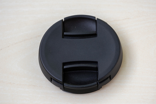 Close-up view of a black camera lens cap without logo on wooden background. A lens cover or lens cap protects camera lenses from scratches, dust, rain, snow, minor collisions, and oily residue.