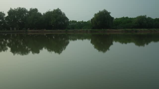 A calm lake with trees in the background. The water of the river is a dark green color. The trees are tall and leafy on a cloudy day in India.
