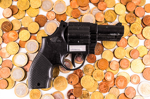 A gun is on top of a pile of coins. The coins are of different sizes and colors, and they are scattered around the gun. Concept of danger and violence, as the gun is a symbol of potential harm