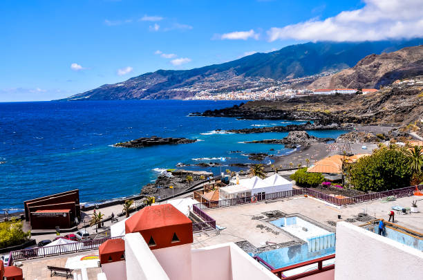 A beautiful beach with a clear blue ocean and a red roof building in the background. The beach is full of people enjoying the day