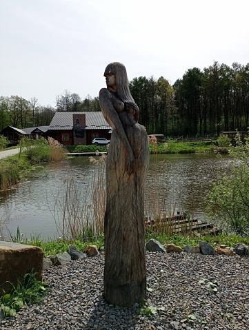 A beautiful wooden sculpture of a young girl against the background of a lake in a park recreation area. The subject of art.