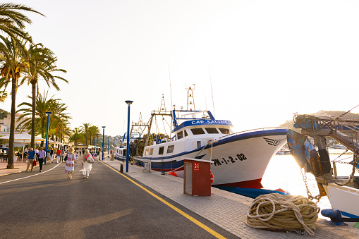 Puerto Andratx, Spain -April 14, 2024:  View of an old fishing boat and tourists walking by the harbor of Andratx in the evening sun. The harbor in Puerto Andratx, Mallorca, is renowned for its affluent international visitors, whether tourists or residents, especially during sunset and for its pub nightlife.