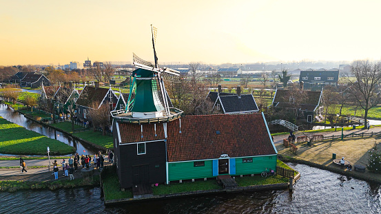 Aerial view of from traditional windmills at Zaanse Schans in the Netherlands, Rotating mills aerial shooting, Rural scene with windmills in Netherlands, Ancient wooden windmills of Zaanse Schans, The most popular tourist destination in the Netherlands

Zaanse Schans is a neighbourhood of Zaandam, near Zaandijk, Netherlands. It is best known for its collection of windmills and wooden houses that were relocated here from the wider region north of Amsterdam for preservation. From 1961 to 1974, old buildings from all over the region known as the Zaanstreek were relocated using lowboy trailers to the Zaanse Schans. Two of the windmills in the Zaanse Schans are preserved in their original site where they were first constructed, and therefore don't make up part of the relocated structures. The Zaans Museum, established in 1994, near the first Zaanse Schans windmill, is located south of the neighbourhood. This architectural reserve for Zaanse timber construction is a protected village scene because of its architectural-historical and landscape value. It developed into an international tourist destination with several million visitors every year: in 2016, there were 1.8 million, in 2017 – 2.2 million.