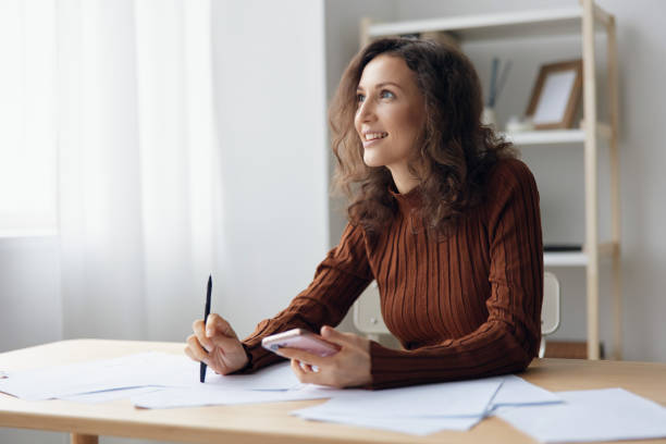 istock Enjoyed laughing happy cheerful curly cute lady sitting at table with papers documents work from home think about future holidays dream about weekend looks aside at window. Copy space 2149013200