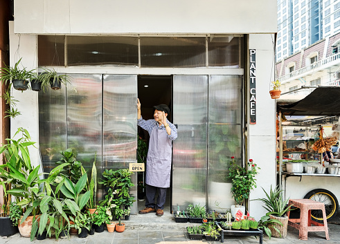 Full length portrait of happy senior waiter standing at coffee shop entrance door looking away and waving hand to someone