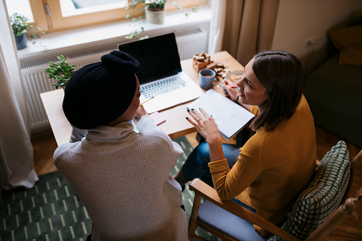 High angle view of two young female business partners working together at home office. Young woman taking notes and discussing with her friend sitting by.