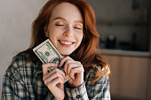 Closeup portrait of happy female freelancer holding in hands wad of dollars that she earned while working remotely at home. Remote earnings, online work.