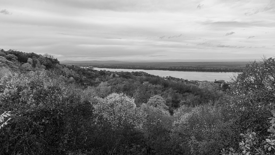 Black and white photo of a wide open view for a high viewpoint of a small town located next to the river surrounded with lush green forest vegetation with a cloudy sky in the background.