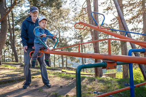 An elderly man supports a young child on a seesaw at a park, creating a scene of inter generational bonding against a forest backdrop with sunlight filtering through trees. Grandpa and grandson enjoying the fun time in the park