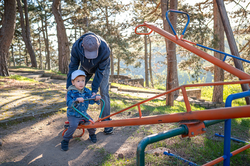 An elderly man supports a young child on a seesaw at a park, creating a scene of inter generational bonding against a forest backdrop with sunlight filtering. through trees. Multiracial 3 years old toddler enjoying his picnic and day out with the family in natural forest like environment