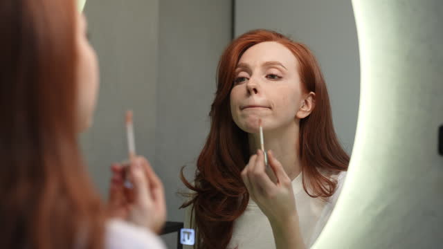 Reflection on mirror of gorgeous young woman applying lipstick get ready preparing for romantic date. Closeup of pretty redhead female model enjoy beauty routine