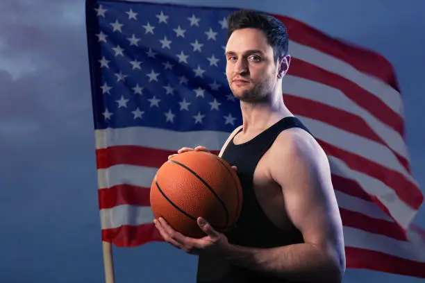 Young white professional basketball player holding a ball in his hands, standing in a heroic pose on the background of the waving USA flag and night sky