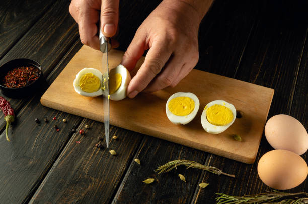 istock Slicing boiled eggs with a knife in the hands of a cook for preparing dinner on the kitchen table. Space for advertising on a dark background 2149024694