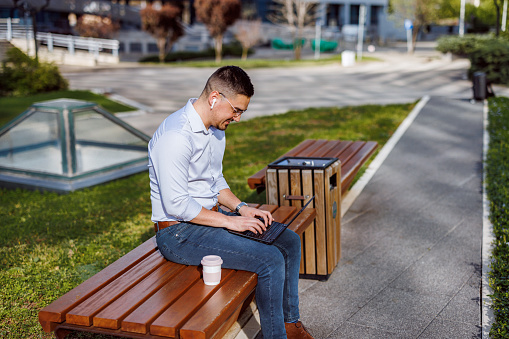 An urban bench becomes a workday retreat for a mid-adult businessman, where he opens his laptop to tackle the challenges of his role within the comforting rhythms of city life
