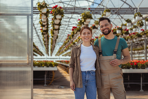 Waist up portrait of smiling farmer with young female worker looking at camera and smiling happily while standing in greenhouse at plantation lit by sunlight