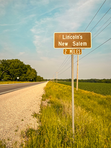 Brown road sign marker on the side of a rural road indicating Abraham Lincoln’s New Salem historic site is two miles away. Agricultural fields and rural country roads in the background of view.