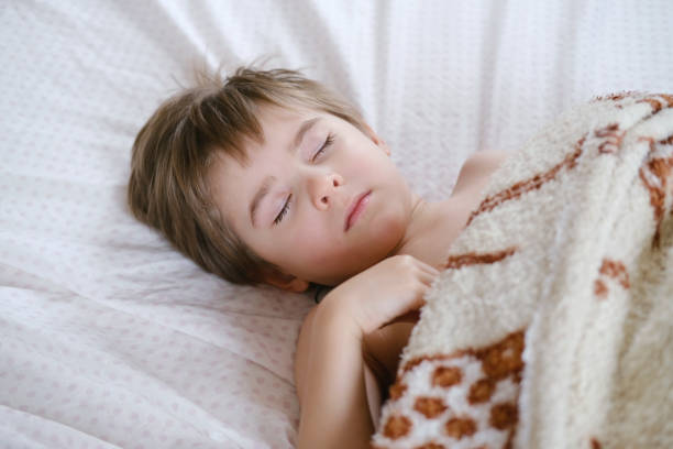 Cute little toddler boy is sleeping with beige plaid at home. Bedtime, napping.