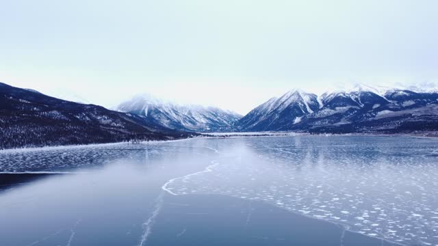 Frozen lake with snow-capped mountains in the background and an overcast sky, dolly