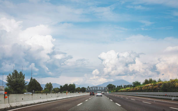 Traveling along the highway between Milan and Venice in Italy, the landscape unfolds beneath a dramatic canopy of clouds. As you journey through the picturesque countryside, the motorway stretches out before you, a ribbon of asphalt cutting through fields and rolling hills. In the distance, the sky is painted with shades of grey, the clouds swirling ominously overhead.