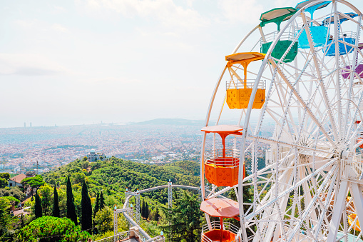 Beathtaking panorama of Barcelona's cityscape and the glistening sea from the heights of Mount Tibidabo and Park Güell and White Ferris wheel turning with colourful baskets against clean sunny blue sky background on sunny day in at Tibidabo, Barcelona