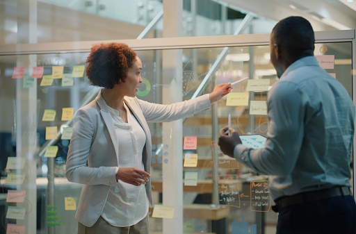 Business people, tablet and sticky notes on glass wall in workplace for planning, discussion and brainstorming. Teamwork, man and woman for sharing ideas, strategy or creative directors in office