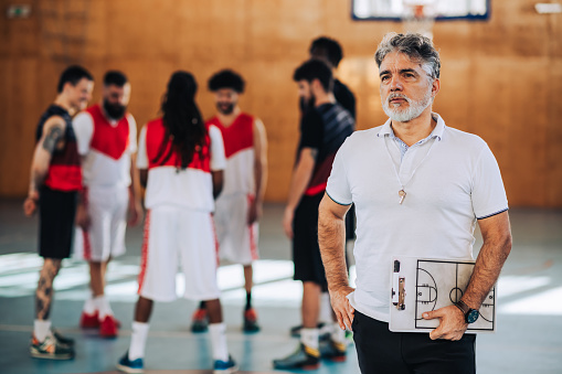 Mature experienced basketball coach holding coach clipboard while standing in court and posing while his team is playing in the background. Portrait of an former athlete training younger players.