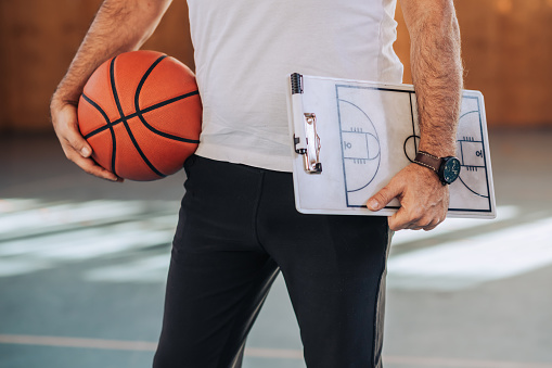 Cropped shot of an unrecognizable elderly caucasian male basketball coach holding a basketball ball and a coach clipboard during a practice in a sports hall. Copy space.