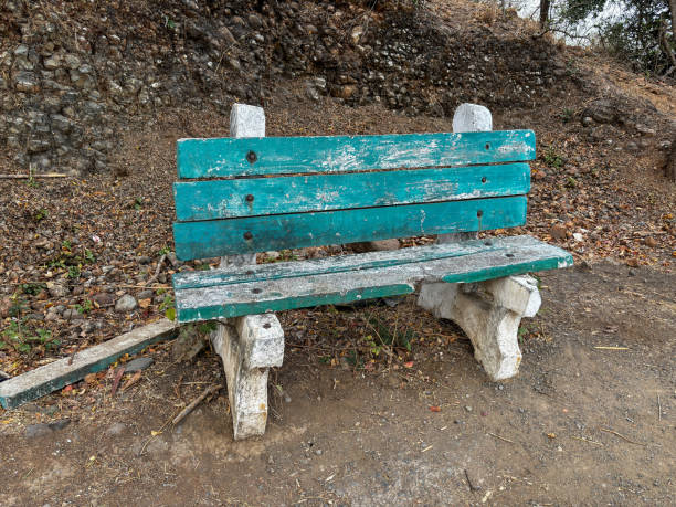 A green bench with a broken back sits on a rocky hillside at Ralamandal, Indore, India. The bench is old and worn, with a few holes in the wood.