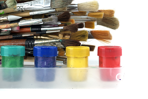 Array of paintbrushes of different sizes and colors alongside cans of acrylic paint with visible colors including red, yellow, blue, and green arranged on a white surface
