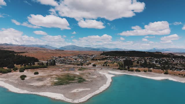 Aerial approaching shot of Lake Tekapo Town in New Zealand with beautiful landscape in summer. Bay, shore and mountain range in background. Panorama wide shot.