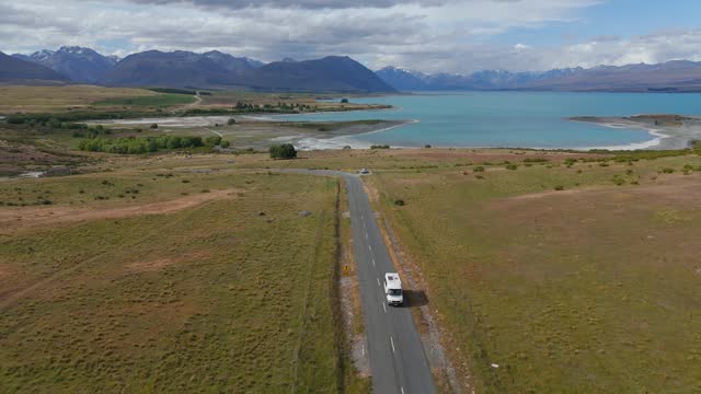 Establishing drone shot of camper van on road near Lake Tekapo in New Zealand. Beautiful scenic fields and snowy mountains in background. Aerial tilt up shot.